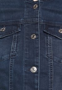 Chaqueta de mezclilla con tejido azul oscuro, herrajes plateados, cierre de botón y bolsillos en el pecho. Textura suave con detalles de costura visibles.