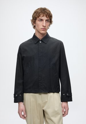 Homme portant une veste noire à col avec des poignets boutonnés et un pantalon plissé beige, debout devant un fond clair uni.