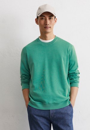 Green cotton crewneck sweatshirt with ribbed cuffs and hem, worn over a white T-shirt. Model wearing denim pants and a beige cap.