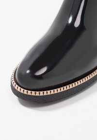 Botte noire brillante à talon, avec une pointe arrondie, dotée d'une semelle noire texturée et d'un bord cousu couleur crème avec des motifs triangulaires.