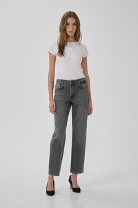 Žena stojaca v bielom krátkom rukáve, s vysokým pásom šedé rovné džínsy a čierne vysoké podpätky na jednoduchom pozadí.