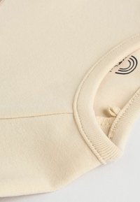 Hautfarbener Baumwollstoff mit glatter Textur, einem gerippten Rand und einem kleinen Logo, das nahe dem Ausschnitt gedruckt ist.