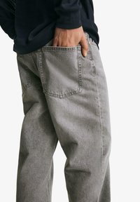 Person wearing gray denim jeans with hand placed inside back pocket, dressed in a black long-sleeve top.