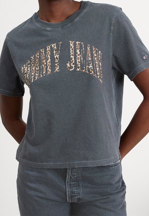 Persoon draagt een gewassen zwarte cropped T-shirt met luipaardprint en de tekst "Tommy Jeans" en bijpassende high-waisted spijkerbroek.