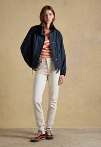 Młoda kobieta stoi, ubrana w granatową kurtkę, pomarańczowo-białą koszulę w paski, kremowe spodnie, białe skarpetki oraz brązowe i czerwone tenisówki.