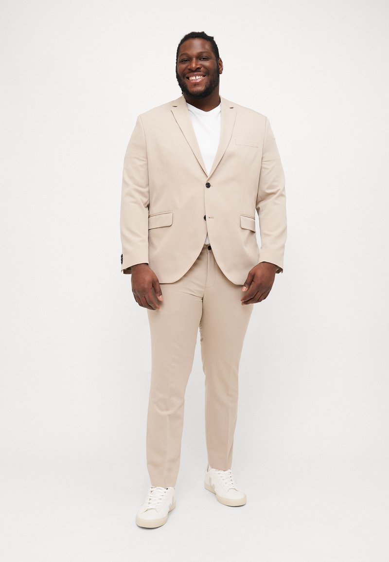 Beige suit with a single-breasted jacket and matching pants, slim-fit design, textured fabric, complemented by white sneakers.