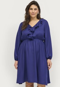 Vestido morado con escote en V, mangas largas y detalles con volantes a lo largo del busto. Tela suave y fluida con una cintura elástica.