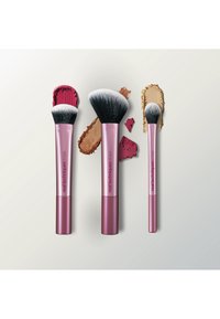 Trois pinceaux de maquillage à manche rose avec des poils noirs et blancs, chacun appliquant différentes teintes de maquillage rouge, marron et beige sur une surface claire.