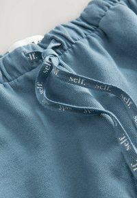 Sinised spordipüksid tekstuurse kangaga, elastne vöökoht ja string, millel on valge sõna "self." Lähedane detail vöökohtast.