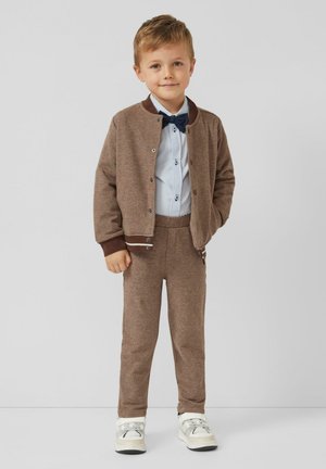 Bruine gebreide cardigan met donkerbruine accenten, lichtblauw overhemd met knopen, bijpassende bruine broek en witte sneakers met grijze details.