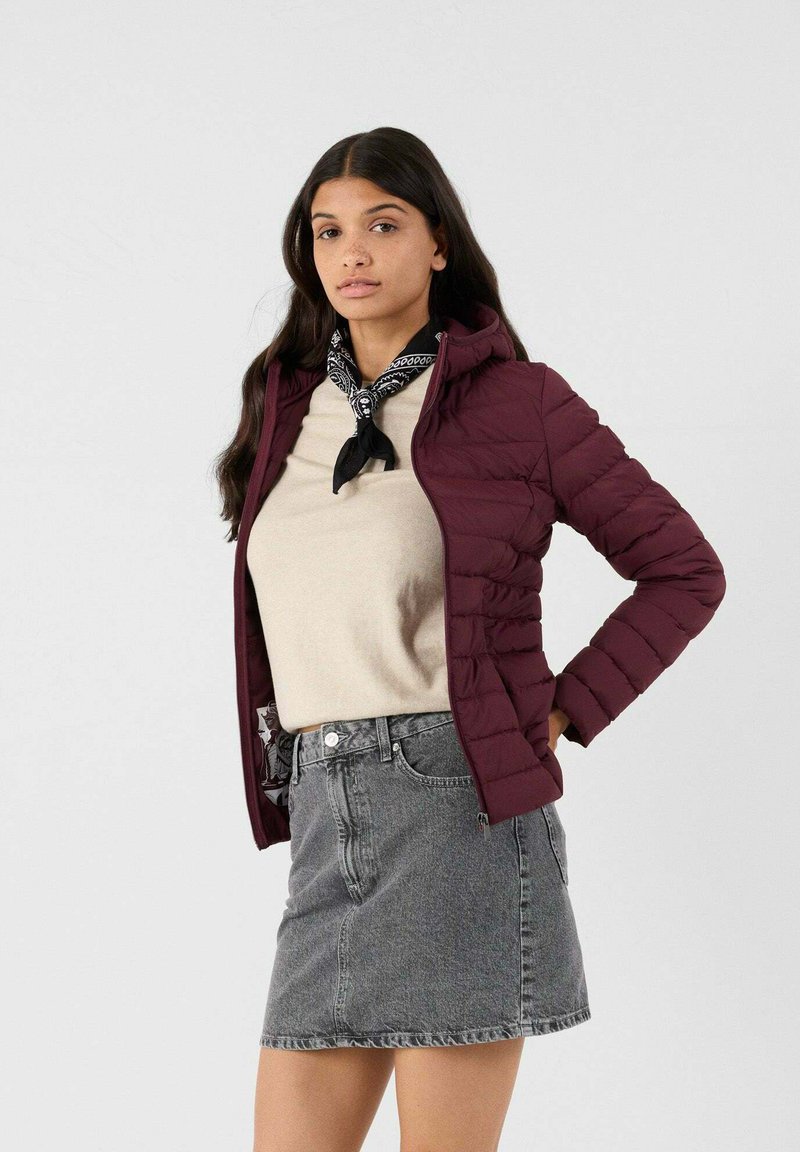 Giacca a vento bordeaux con cappuccio, design trapuntato, indossata sopra un top beige e una gonna di denim grigia. Sciarpa a scacchi bianca e nera annodata al collo.