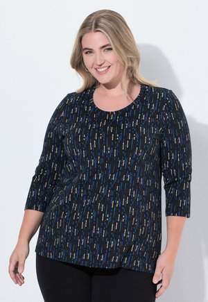 Usměvavá žena s blond vlasy oblečená v černém topu s dlouhým rukávem, který má modré a béžové svislé čárkované vzory, a v černých kalhotách.