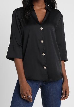Vrouw draagt een zwarte blouse met knopen en driekwart mouwen en blauwe spijkerbroek, toont ringen aan beide handen.