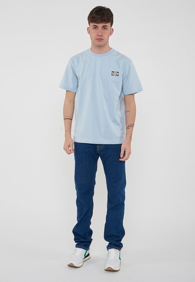 Camiseta de algodón azul claro con mangas cortas, escote redondo y un pequeño logo impreso en el lado izquierdo del pecho. Combinada con unos vaqueros azul oscuro y zapatillas blancas.