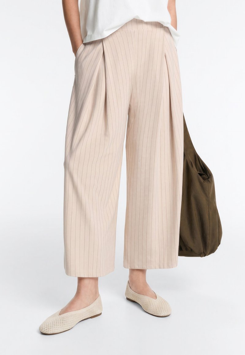 Pantalon beige à jambes larges rayé fin, raccourci, associé à des ballerines tissées crème et un haut blanc, tenant un sac cabas vert olive foncé.