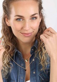 Gouden hartvormige colliers, één groter en één kleiner, gedragen over een denim shirt. De gestructureerde ketting voegt detail toe; beide colliers hangen op verschillende lengtes.