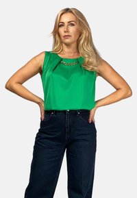 Zielony top bez rękawów z fałdą z przodu i złotym łańcuchem, w połączeniu z wysokim stanem ciemnoniebieskimi dżinsami. Gładka tekstura, luźny krój.