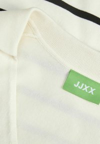Balta megztinė medžiaga su juodomis juostelėmis, su giliu apykakle ir žaliu etiketu "JJXX". Minkšta tekstūra ir laisvalaikio dizainas.
