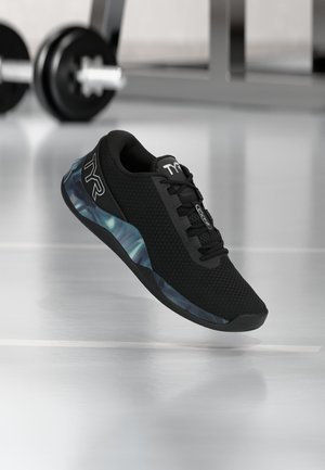 Zapatilla deportiva negra con detalle de suela azul con patrón, flotando sobre un suelo brillante de gimnasio con pesas desenfocadas en el fondo.