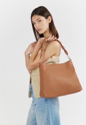 Giovane donna con top beige e jeans azzurri tiene una grande borsa marrone in pelle con manici intrecciati sulla spalla.