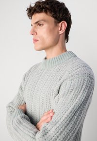 Young man with curly hair wearing a light grey textured sweater, arms crossed, looking to the left against a plain background.