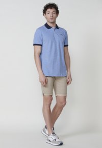 Gaiši zils polo krekls ar teksturētu audumu, tumši zila apkakle un piedurknes, apvienots ar bēšiem šortiem. Baltas kedas ar pelēkiem akcentiem.