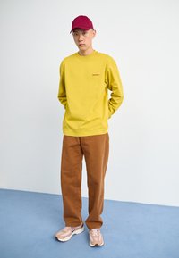 Yellow long-sleeve shirt with logo, brown pants, and light pink sneakers. Model wears a burgundy cap, hands in pockets, standing against a plain background.
