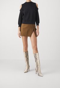 Mujer vistiendo una blusa negra de hombros descubiertos, una falda corta marrón con abertura frontal y botas de tacón de serpiente hasta la rodilla.