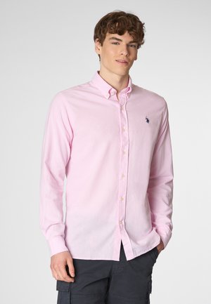 Giovane uomo con capelli ricci che indossa una camicia rosa con bottoni, in piedi davanti a uno sfondo neutro, sorridendo con sicurezza.