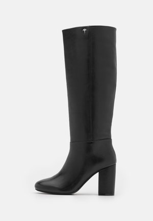 Czarny, sięgający kolan, skórzany kozak na słupku z małym metalowym logo w pobliżu góry na jasnym tle.