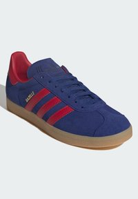 Blå ruskinds Adidas Gazelle sneaker med røde striber og hælklap, beige struktureret sål, og "GAZELLE" i guldtekst på siden.