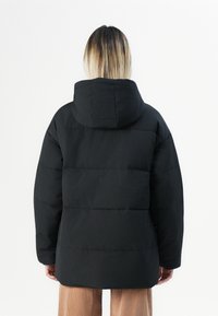 Person med skulderlangt blondt hår iført en svart hettejakke i puff-stil og beige bukser, vendt mot en hvit bakgrunn.