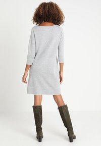 Una donna con i capelli ricci è in piedi di spalle indossando un vestito grigio chiaro fino al ginocchio e stivali a tacco alto color verde scuro fino al ginocchio.