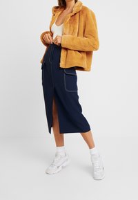 Kvinna som bär en senapsgul fuskpälsjacka, vit linne, marinblå midi-kjol med framslits och stora sidofickor, vita sneakers med strumpor.