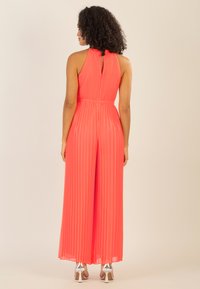 Mono de tirantes plisado color coral con cuello alto, cintura elástica y un detalle de ojal en la parte posterior, que presenta piernas anchas y zapatos de tacón alto metálicos.