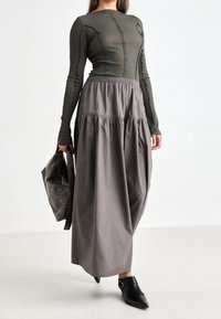 Long-sleeve, fitted green top paired with a flowing, gray maxi skirt featuring gathered waist and textured fabric. Model holds a dark bag.