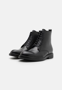 Botine negre din piele cu un finisaj neted, design cu șireturi, vârf rotunjit și talpă din cauciuc cu profil scăzut. Au un tab de apăsare pe spate.