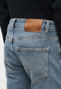 Κοντινή λήψη ατόμου που φοράει ανοιχτό μπλε τζιν με ραμμένη πίσω τσέπη και καφέ δερμάτινο ταμπελάκι της μάρκας στη μέση, συνδυασμένο με μαύρο λαστιχωτό τοπ.