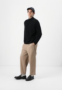 Maglione nero, pantaloni beige e scarpe nere. Il modello indossa un berretto grigio. Il maglione ha un collo alto; i pantaloni hanno una vestibilità rilassata.