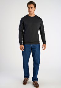 Felpa nera con collo rotondo, maniche lunghe e polsini a costine, abbinata a jeans blu e scarpe da barca marroni su uno sfondo neutro.