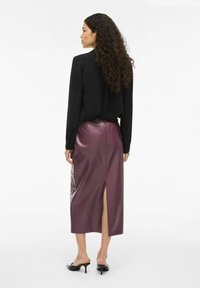 Schwarze Bluse mit langen Ärmeln, kombiniert mit einem lila Leder-Midirock mit einem Schlitz hinten. Das Model trägt schwarze offene High Heels.