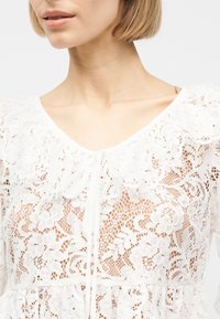 Femme portant un chemisier en dentelle florale blanche avec col festonné et encolure en V, montrant le cou et une partie de ses cheveux blonds courts.