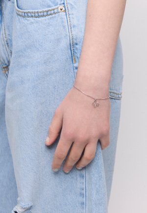 Srebrna bransoletka łańcuszkowa z małym otwartym wisiorkiem krzyża, noszona na ręce na tle jasnoniebieskiego dżinsu; gładka faktura, delikatny design.