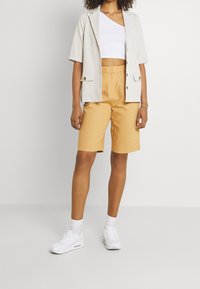 Blazer beige a maniche corte sopra un crop top bianco, abbinato a pantaloni corti senape. Il modello indossa sneakers bianche e calzini bianchi. Design e texture semplici.