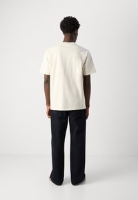 Tricou scurt cu mânecă, de culoare crem, confecționat din bumbac, purtat cu pantaloni largi de culoare albastră închis și încălțăminte casual, văzut din spate.