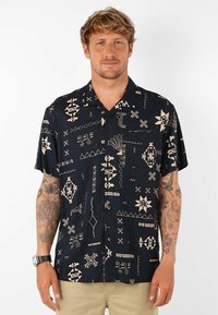 Chemise à manches courtes noire avec des motifs géométriques et abstraits beige, fabriquée en tissu léger, présentant une coupe décontractée et un devant boutonné.