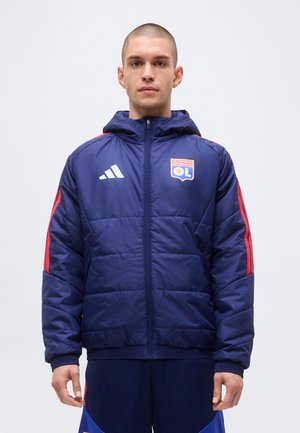 Jeune homme portant une veste matelassée bleue Adidas avec le logo de l'Olympique Lyonnais et des bandes rouges sur les manches, debout devant un fond uni.