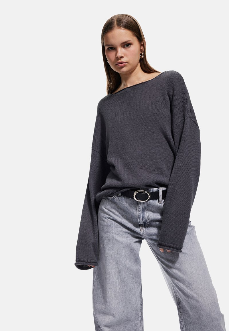 Übergroßer grauer Pullover mit gerippter Textur, weiten Ärmeln und einem weitem Rundhalsausschnitt, kombiniert mit hellgrauen Jeans und einem schwarzen Gürtel.