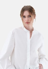 Camicia bianca in cotone a maniche lunghe con colletto rialzato, caratterizzata da una chiusura con bottoni e una vestibilità rilassata, presentata su uno sfondo chiaro.