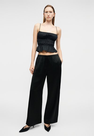 Νεαρή γυναίκα που φοράει μαύρο κοντό τοπ με ρεγκλάν, φαρδιά μαύρη παντελόνα και μαύρες μυτερές γόβες, στέκεται μπροστά σε απλό φωτεινό φόντο.